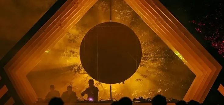 Escenario con un gran círculo de sol y un DJ entre humo dorado